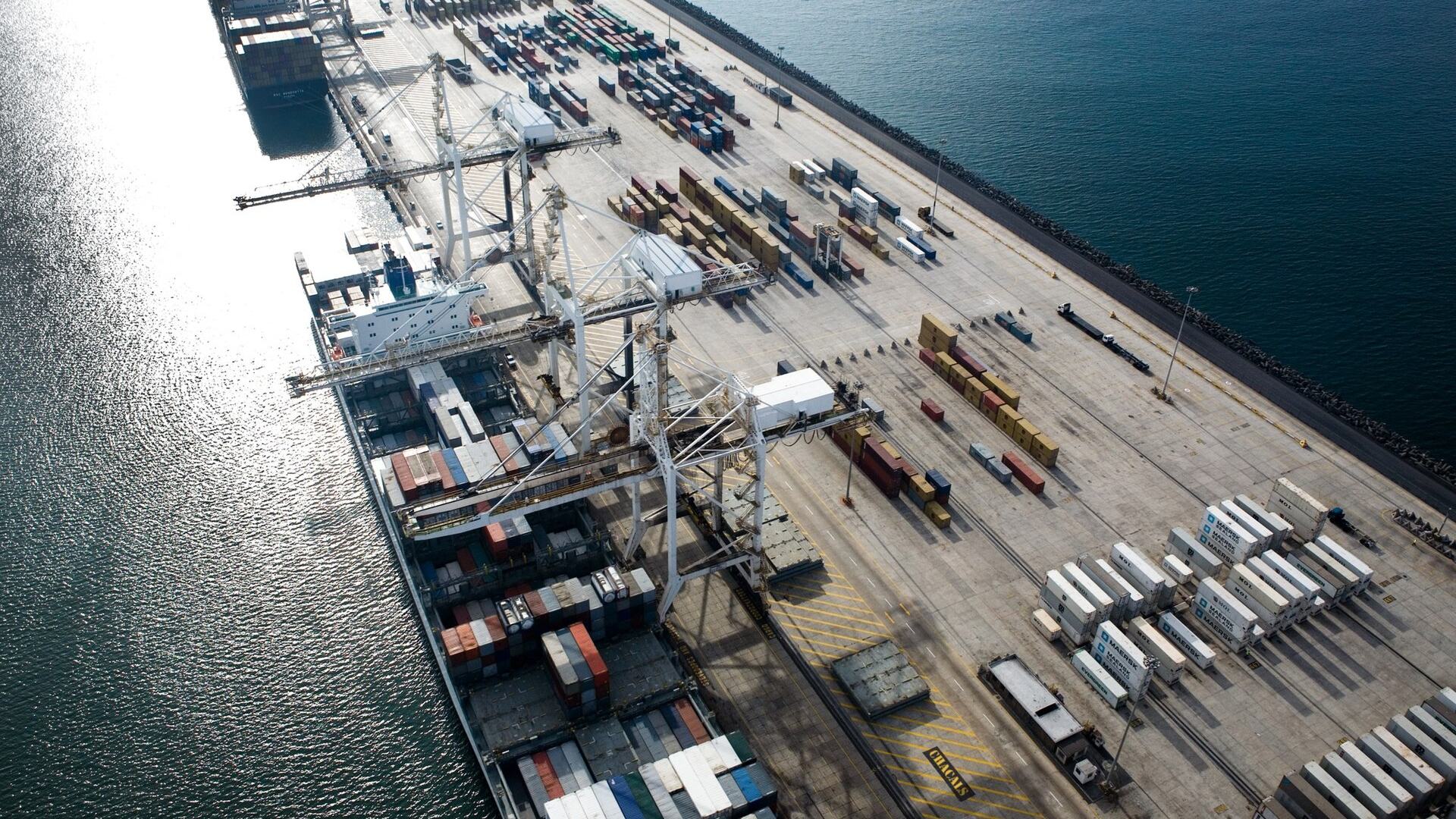 Aerial view of a container terminal: a ship at berth beneath gantry cranes, containers stacked the length of the quay.